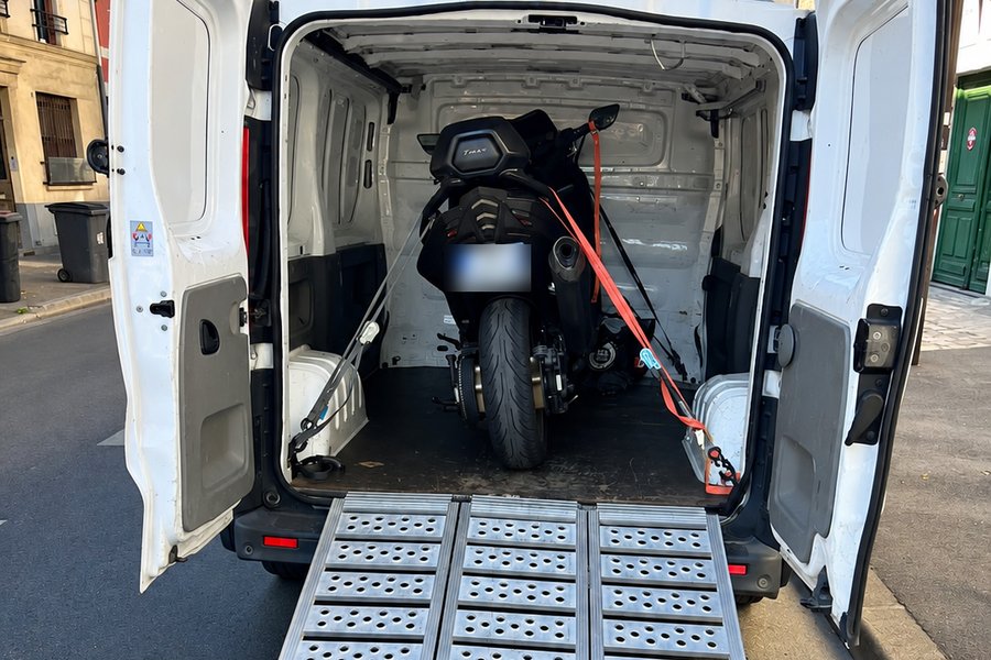 Camion fermé portes ouvertes, rampe d’accès dépliée sur la chaussée, scooter sanglé à l’intérieur